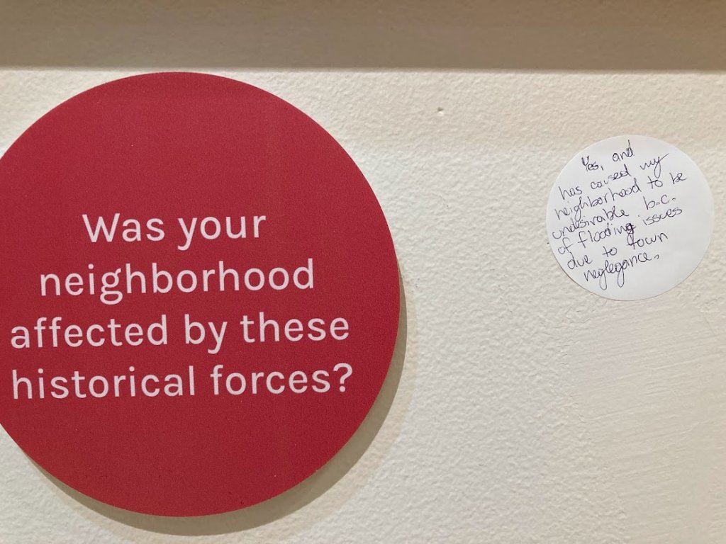 Content from the exhibition hangs on a wall: A prompt asks "was your neighborhood affected by these historical forces?" A sticky-note replies "Yes and it has caused my neighborhood to become undesirable due to flooding issues caused by negligence."